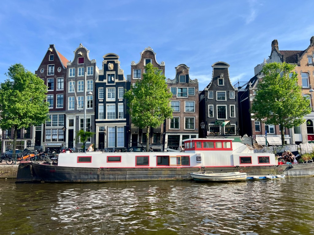 A white houseboat with red trim moored along an Amsterdam canal in front of narrow gabled houses.